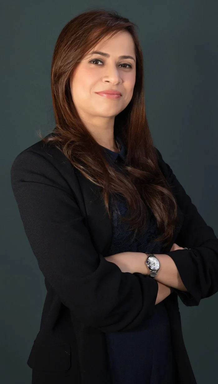 A woman with long brown hair, wearing a black blazer over a dark top, standing with her arms crossed against a dark gray background.