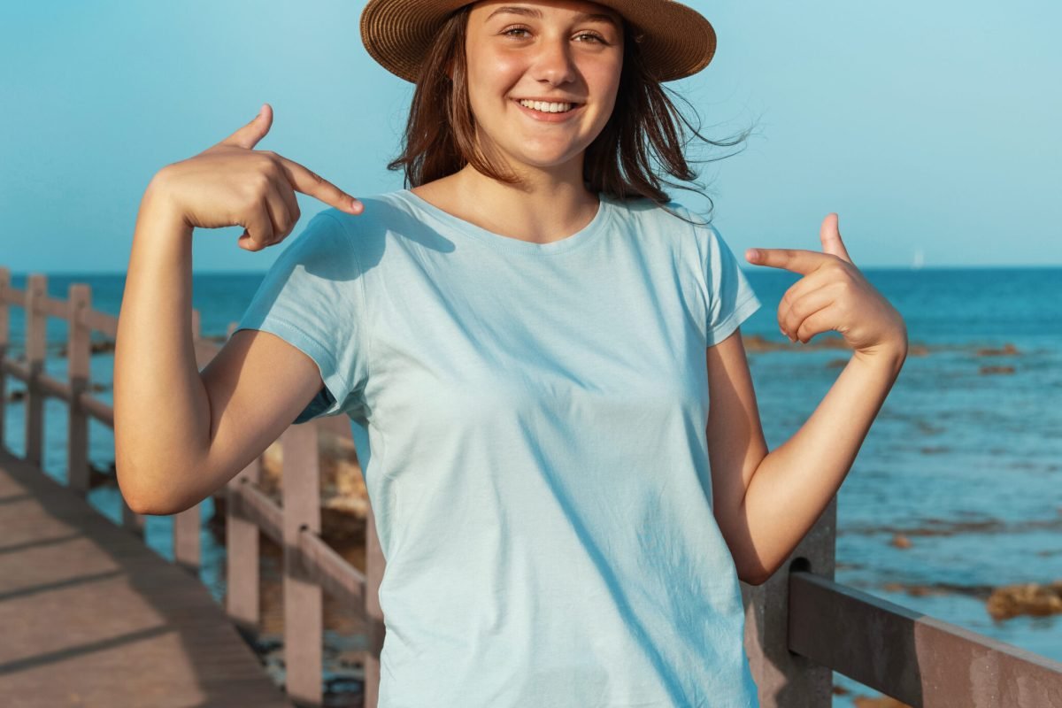 Smiling teenage girl staying on wooden sidewalk by the sea wearing straw hat, light blue t-shirt and pointing at it. T-shirt mockup