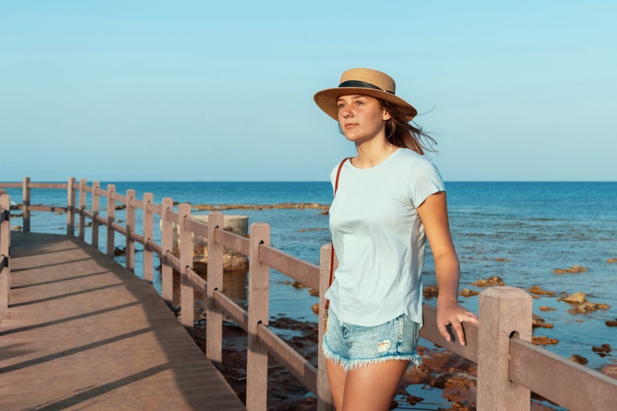 Teen girl standing on wooden walkway by the sea at sunset, wearing light blue t-shirt, straw hat and purse. Summer travel concept
